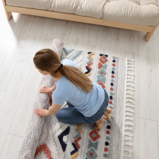 Illustration - Ihr Experte für schönes Wohnen - Frau rollt Teppich mit schönem Muster auf dem Boden im Zimmer aus, Blick von oben.